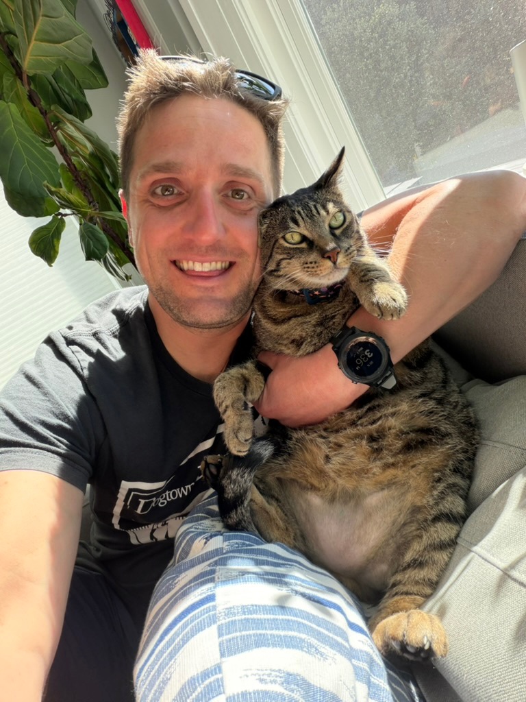 J.J. smiling and holding his cat Baxter by a sunny window